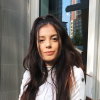 Woman with long dark hair, wearing a white top, against a blurred background.