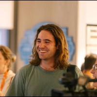 Smiling man with shoulder length light brown hair, wearing a casual green shirt, in an indoor setting.