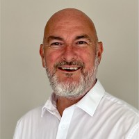 Smiling bald man with a white beard, wearing a white shirt, against a neutral background.