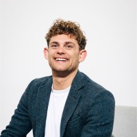 Smiling man with curly hair, wearing a blazer and white shirt, against a light background.